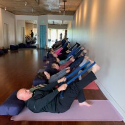 Mixed age class demonstrates Avita Yoga lying supine with strap around right foot and leg extended in the air.