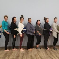 Nine women with yoga blankets belted to their bellies to simulate a pregnant belly in JAI Prenatal Yoga for Birthing teacher training.